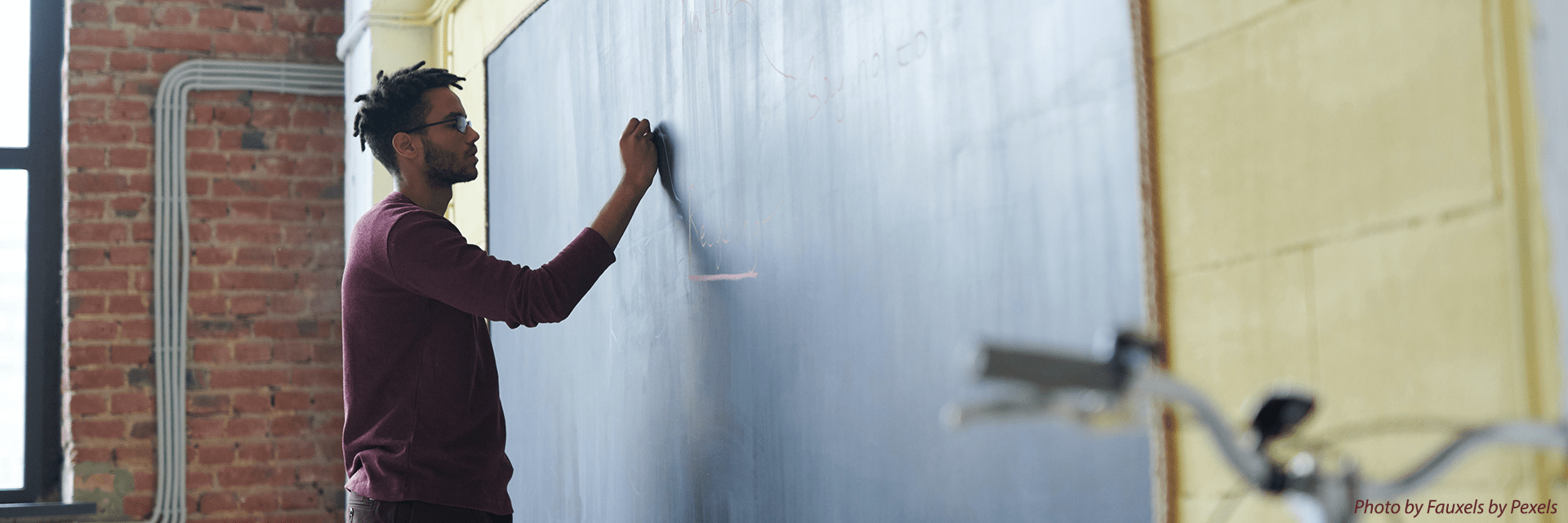 A black male in a burgandy sweater stands at the chalkboard writing words as he teaches a class.